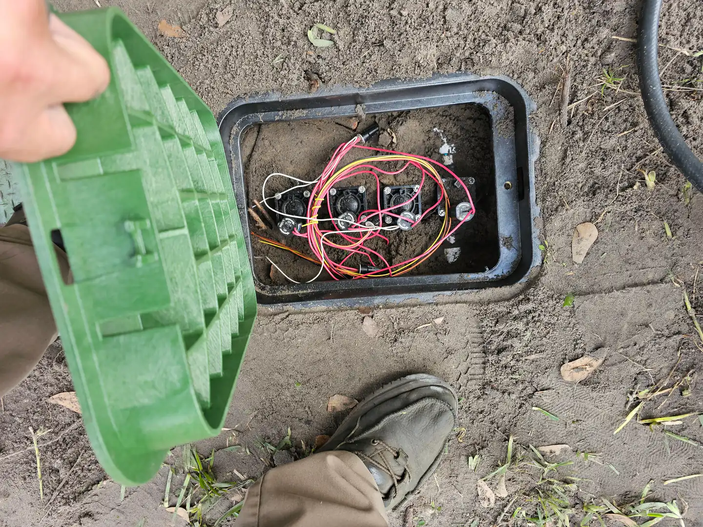 A hand lifting the lid off an in-ground irrigation valve box, showing three solenoid valves and a nest of red and yellow wiring, with a work boot on the ground beside it