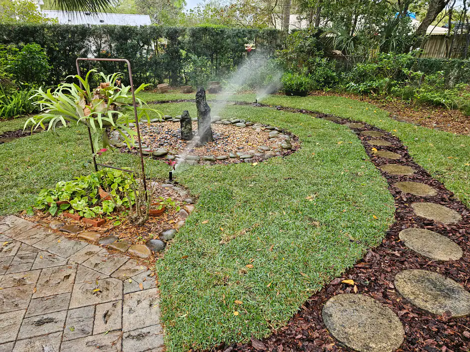 A Woods irrigation system running - sprinklers throwing an even arc over a lush backyard with a circular stone garden and fresh sod